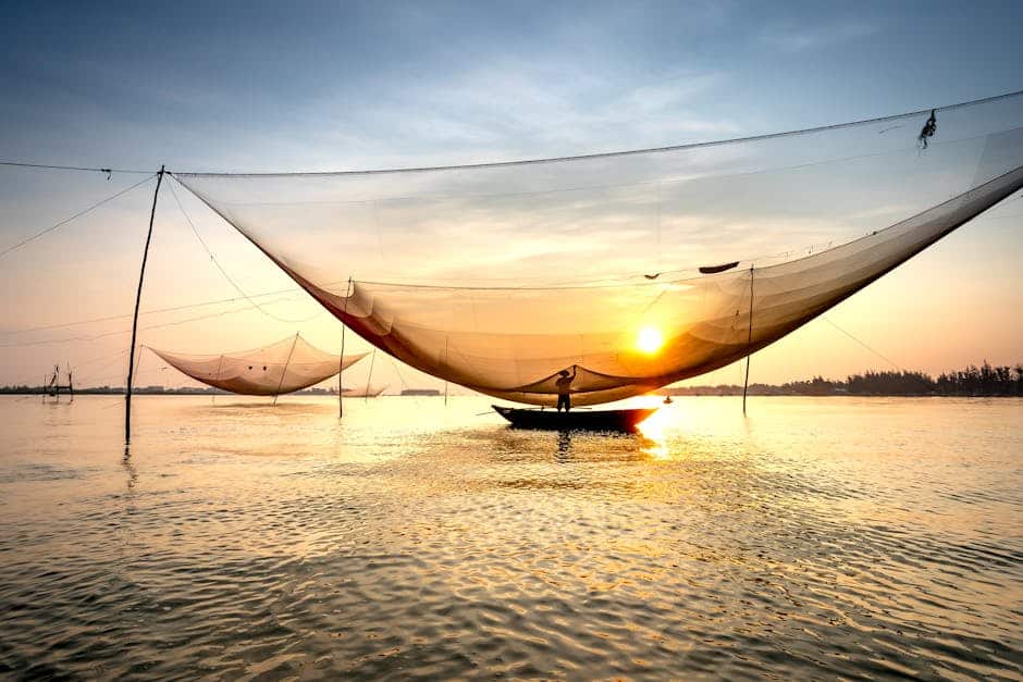 Fishing boat with large net is illuminated by the setting sun on a calm body of water. The sky is a blend of blue and orange hues.